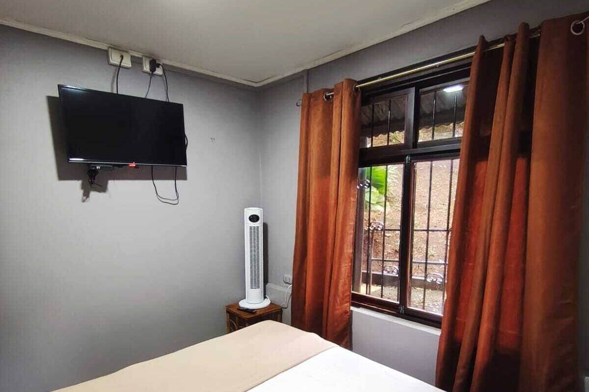 A room with a bed, a TV mounted on the wall, an air conditioner, and a window with curtains.