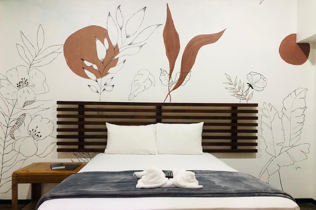 A hotel room with a bed, wooden headboard, white pillows, a nightstand with a book, and a wall with floral and leaf patterns.