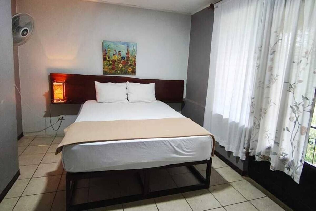 A hotel room with a bed, a painting on the wall, a window with curtains, and a ceiling fan.