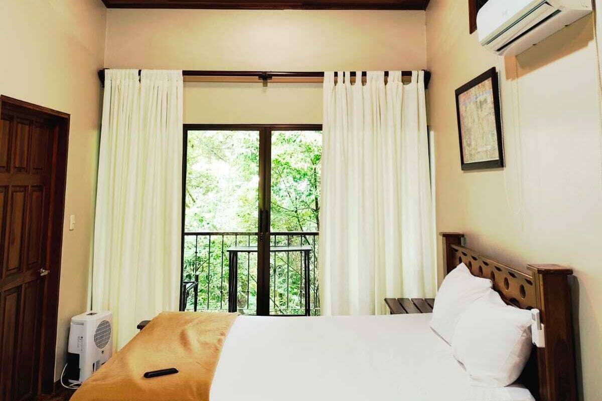 A bedroom with a wooden ceiling, a bed with white bedding, a window with curtains, and an air conditioning unit.