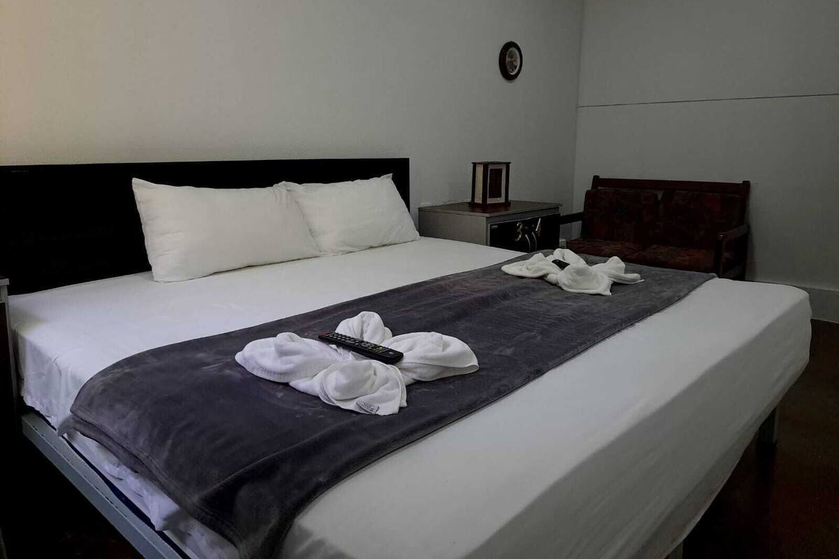 A neatly made bed with white linens, a dark grey blanket, and white towels folded on the bed. There is a remote control on the blanket.