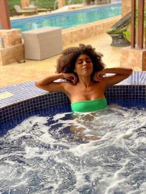 Une personne se trouve dans un jacuzzi dont les bords sont recouverts de carreaux, entouré d’un espace piscine comprenant des chaises longues et un toboggan.