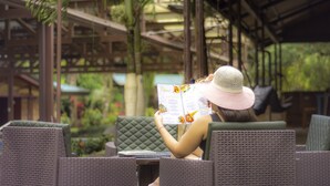 A person reading a menu in a covered outdoor seating area with wicker furniture and palm trees in the background. - Avita Lodge Hotel de Montaña (Quesada)