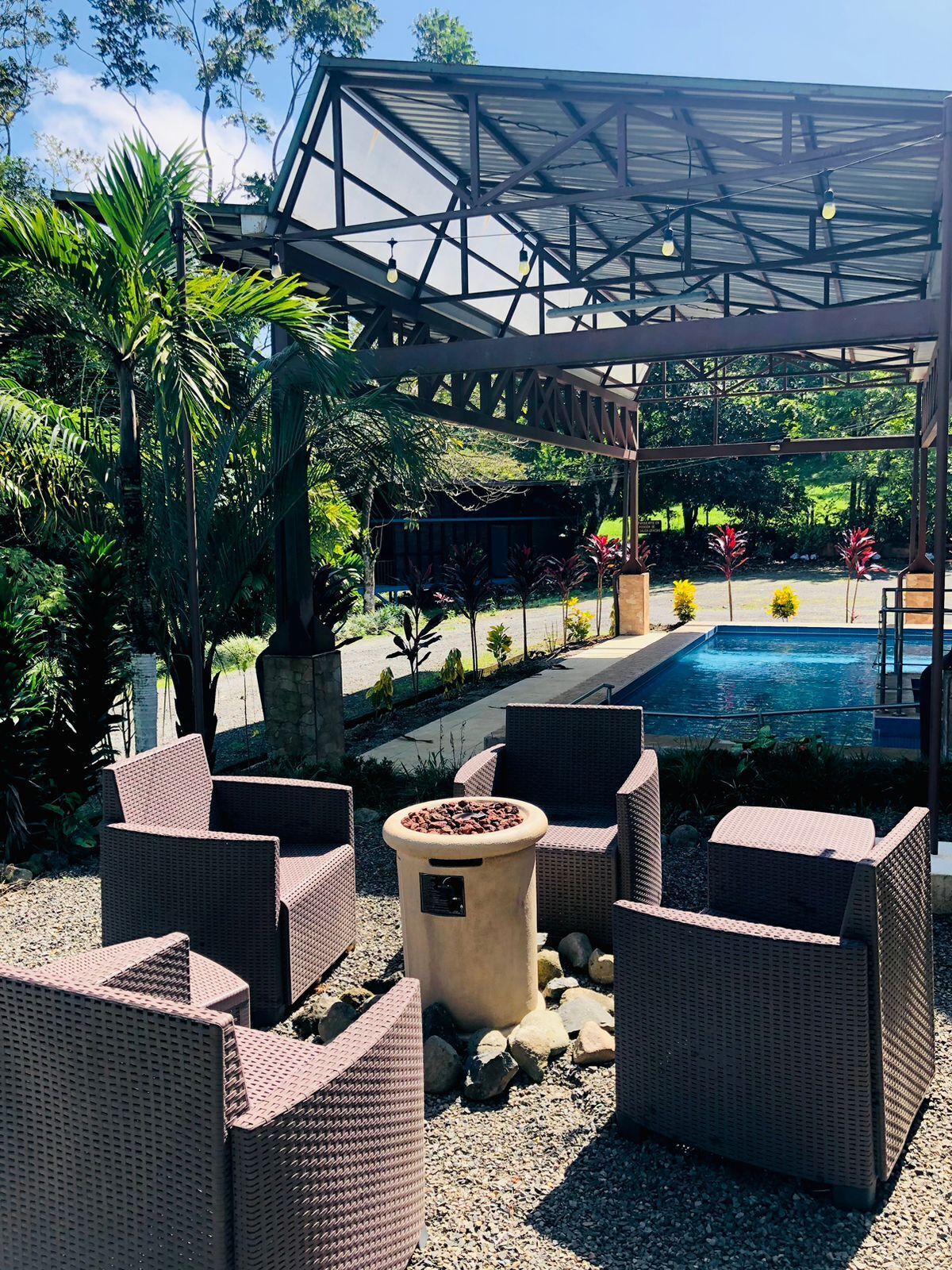 Un espace extérieur aménagé avec des chaises en osier et un foyer, entouré d’une piscine et d’une végétation luxuriante.