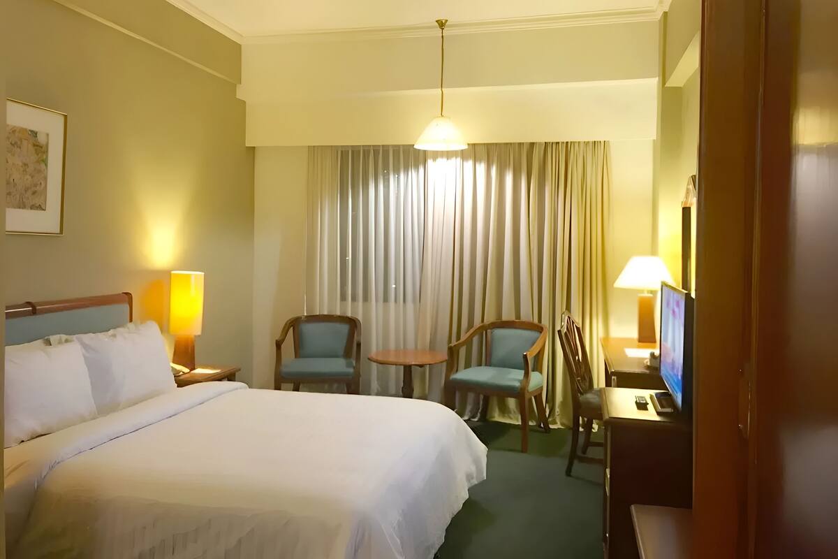 A hotel room with a large bed, two chairs, a small table, a TV, and a window with curtains.