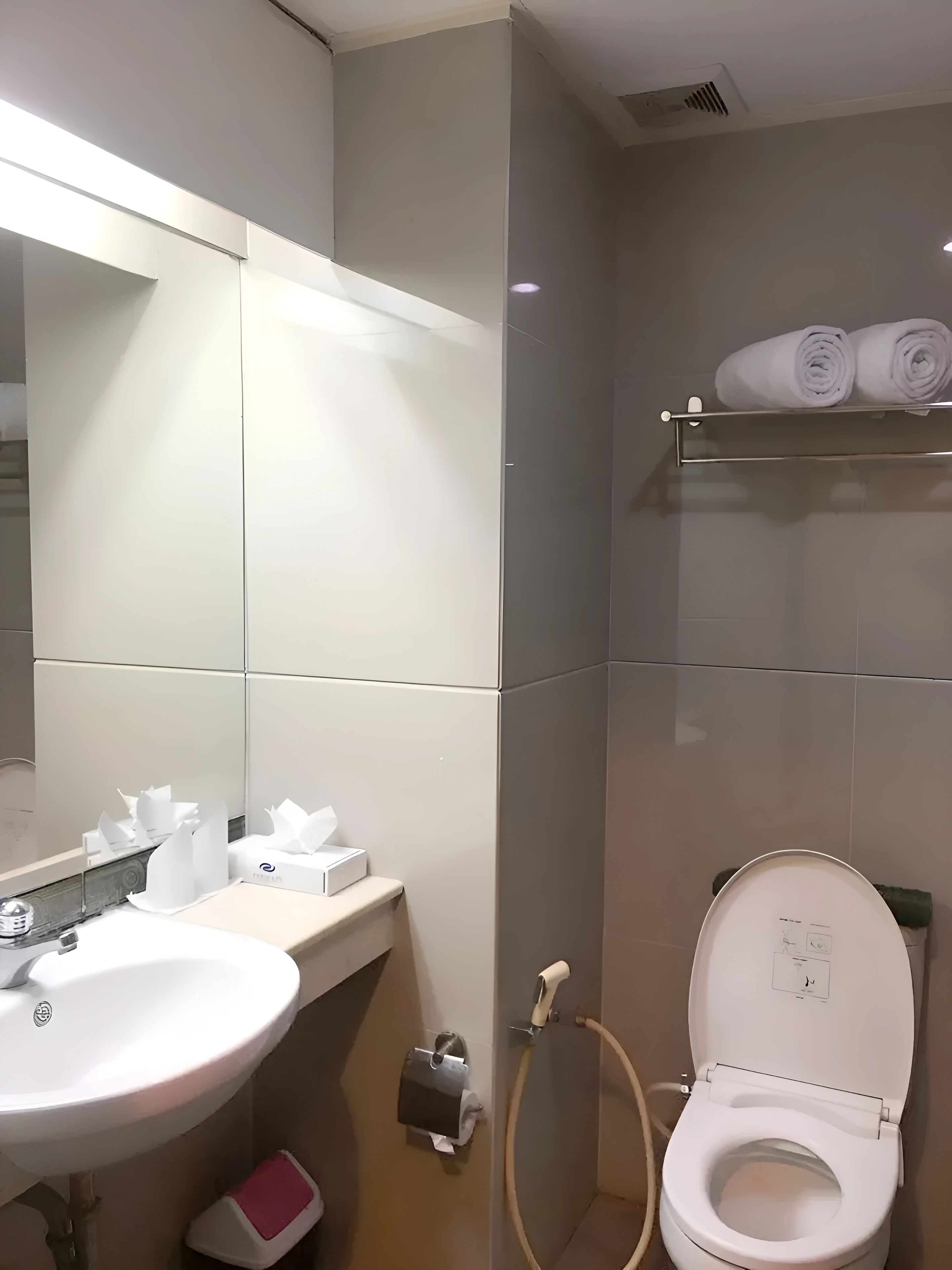 Un baño moderno con un lavamanos blanco, espejo y inodoro. Hay toallas en un estante y un dispensador de papel higiénico en la pared.