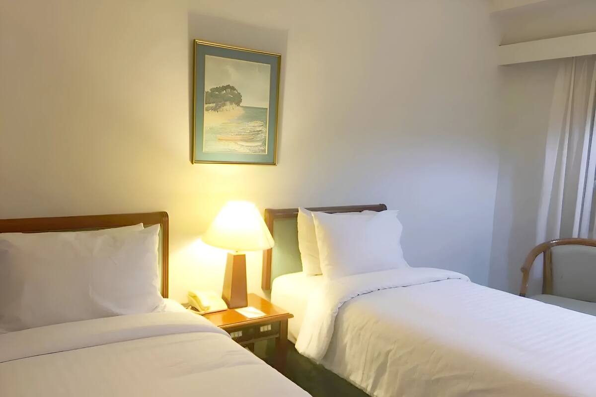 A hotel room with two beds, a bedside table with a lamp, and a framed picture on the wall.