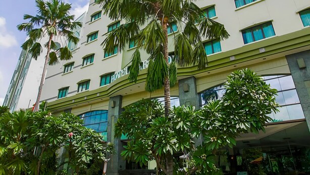 Un hotel de varios pisos con diseño moderno, que presenta una entrada central con un paseo techado, rodeado de abundante vegetación y palmeras.