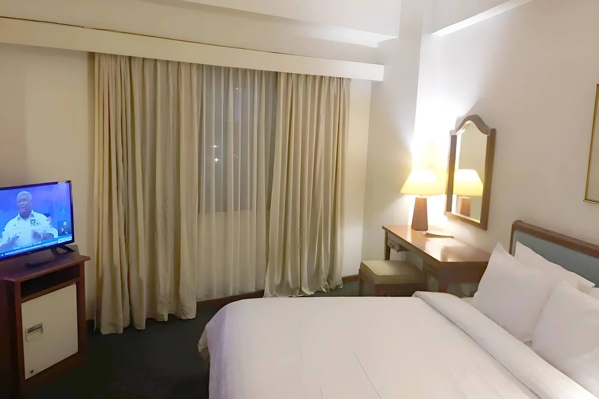 A hotel room with a bed, a desk with a lamp, a television, and a large window with curtains.