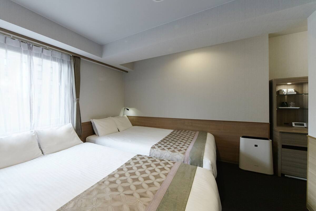 A hotel room with two beds, a bedside table with a lamp, a small desk, and a trash bin.