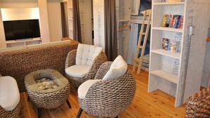 A living room with a large flat-screen TV, a sofa, two armchairs, a coffee table, and a bookshelf with magazines.