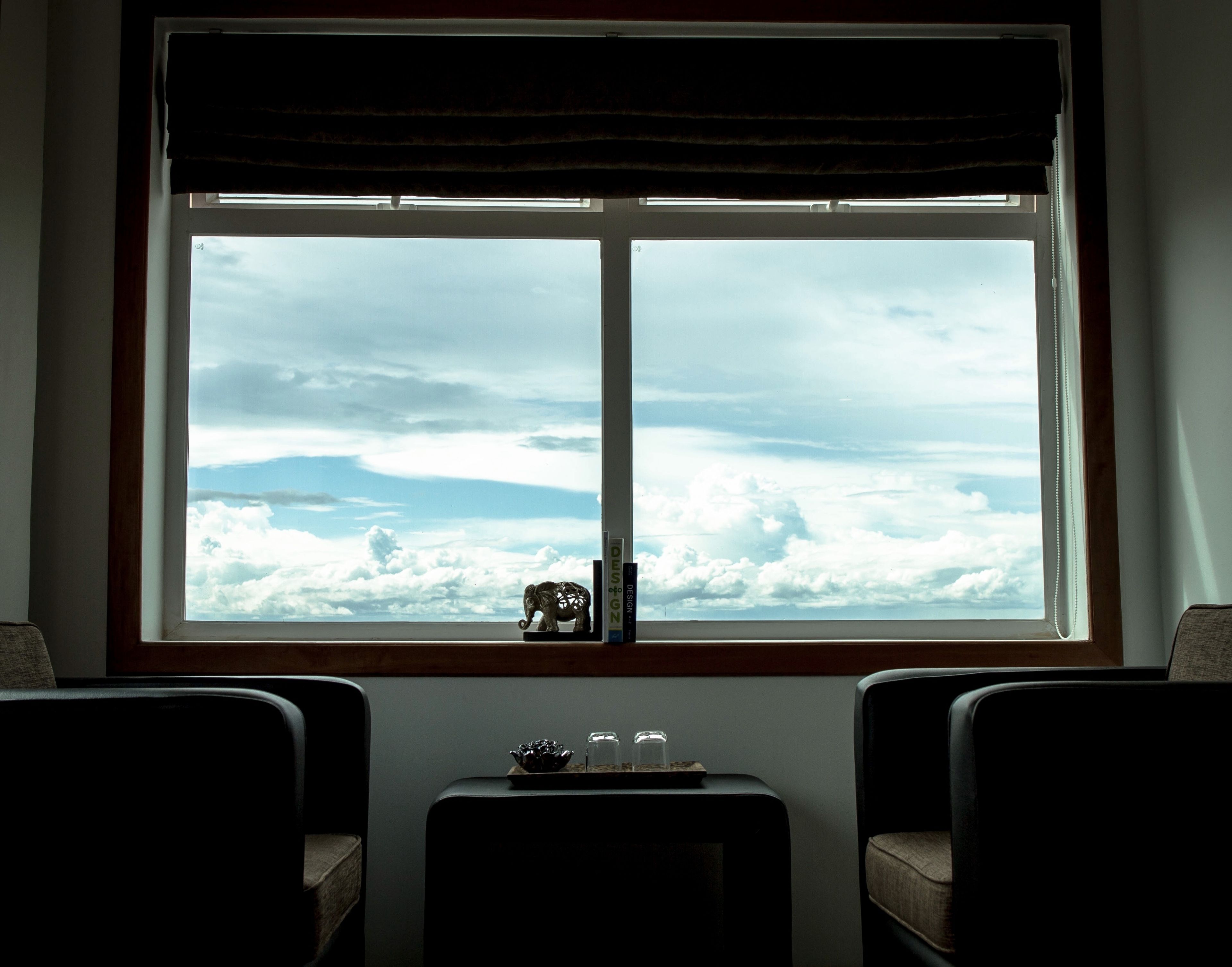 A room with a large window overlooking clouds and mountains, a small table with decorative items, and two chairs.