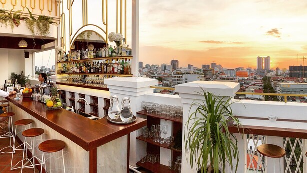 A rooftop bar with a city skyline view, bar stools, shelves with bottles and glasses, and a decorative plant. - Point Boutique Hotel (Phnom Penh)