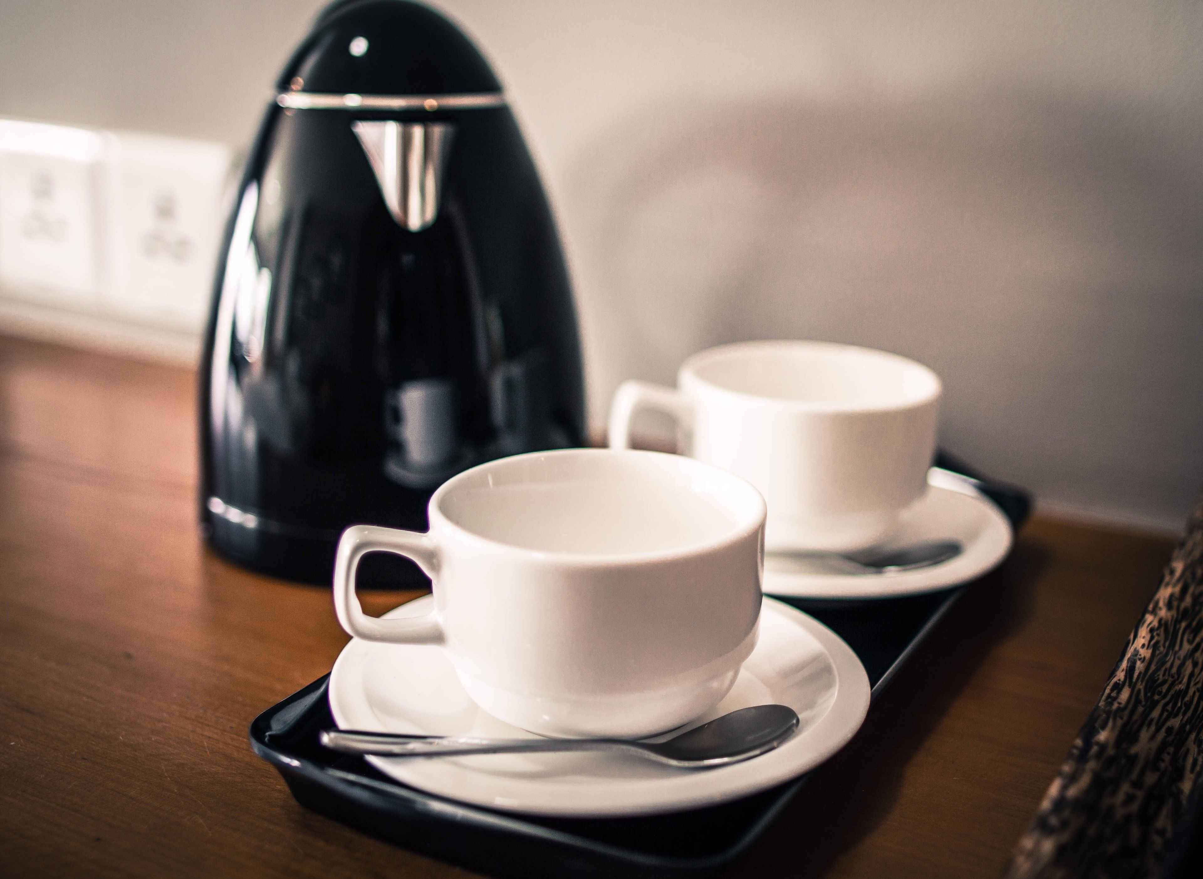 Two white cups on saucers with a spoon, a black coffee maker, and a tray on a wooden table.