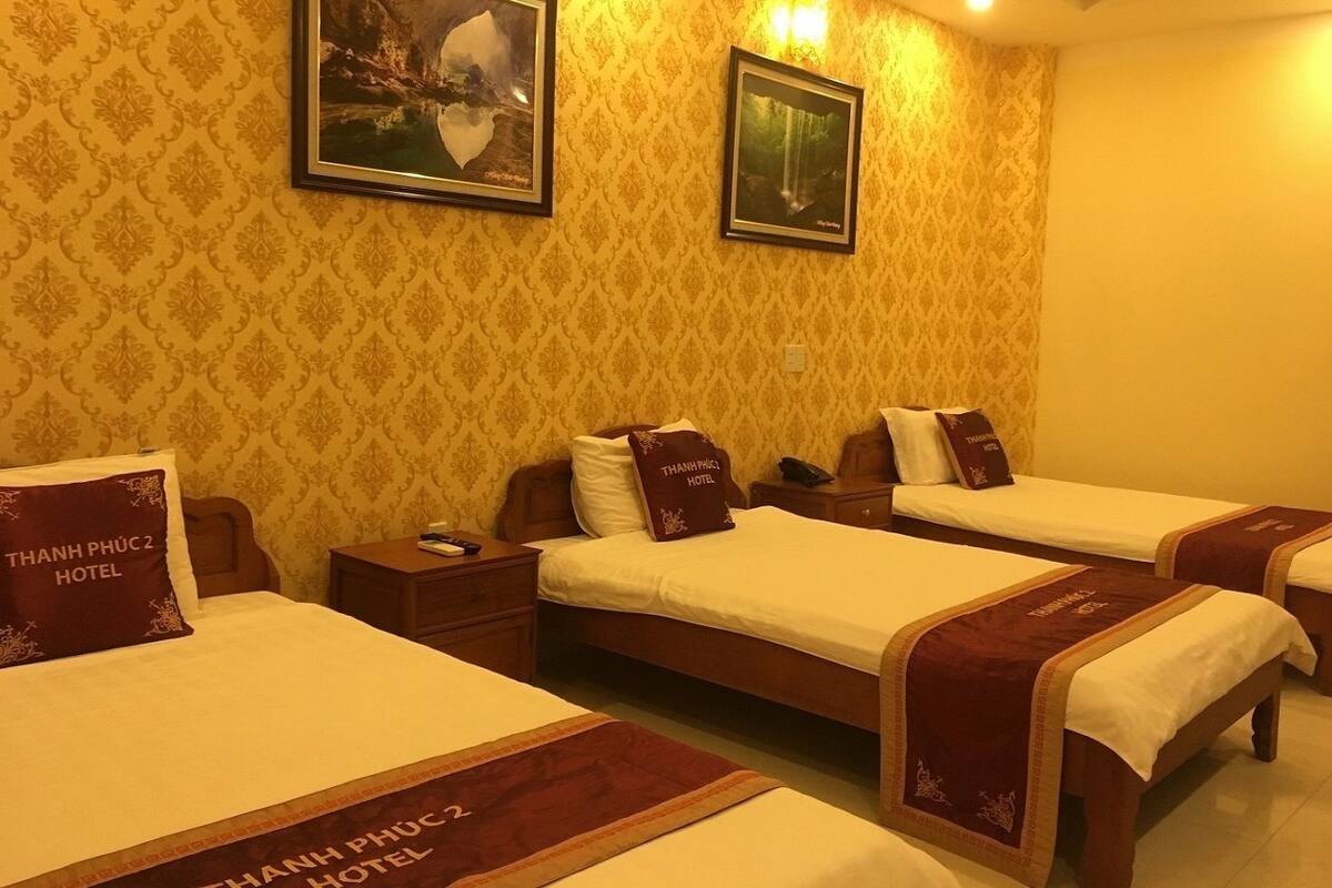 Habitación de hotel con tres camas, cada una con almohada y cubrecama con el logotipo 'THANH PHU C2 HOTEL'. En la pared hay dos cuadros enmarcados, una mesita de noche con un teléfono y una lámpara.