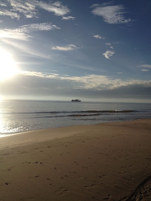A beach with footprints leading to the water, a ship in the distance, and a clear sky.