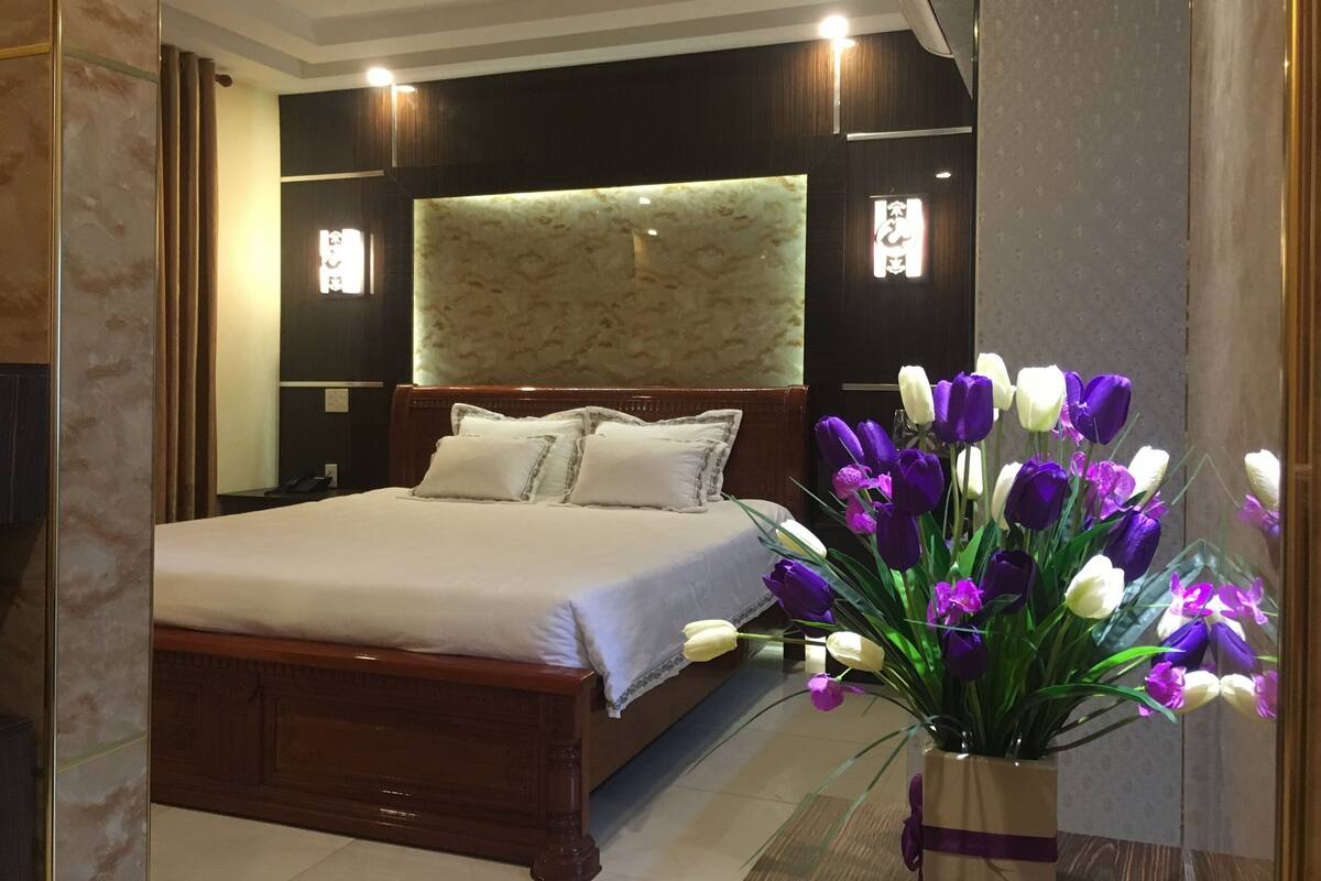 Habitación de hotel con una cama grande, un cabecero de madera y un florero con flores moradas y blancas.
