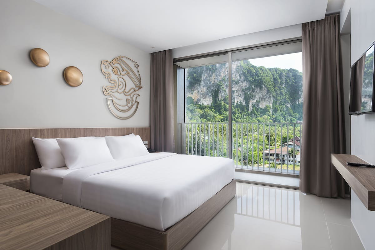 A modern hotel room with a large bed, a wooden nightstand, a flat-screen TV, and a balcony with a view of a mountainous landscape.