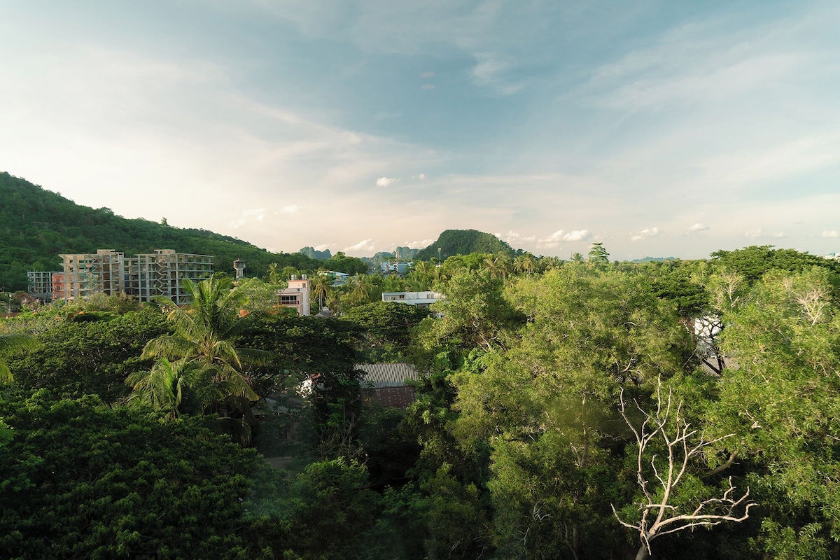 A view of a building surrounded by dense greenery and hills in the background.