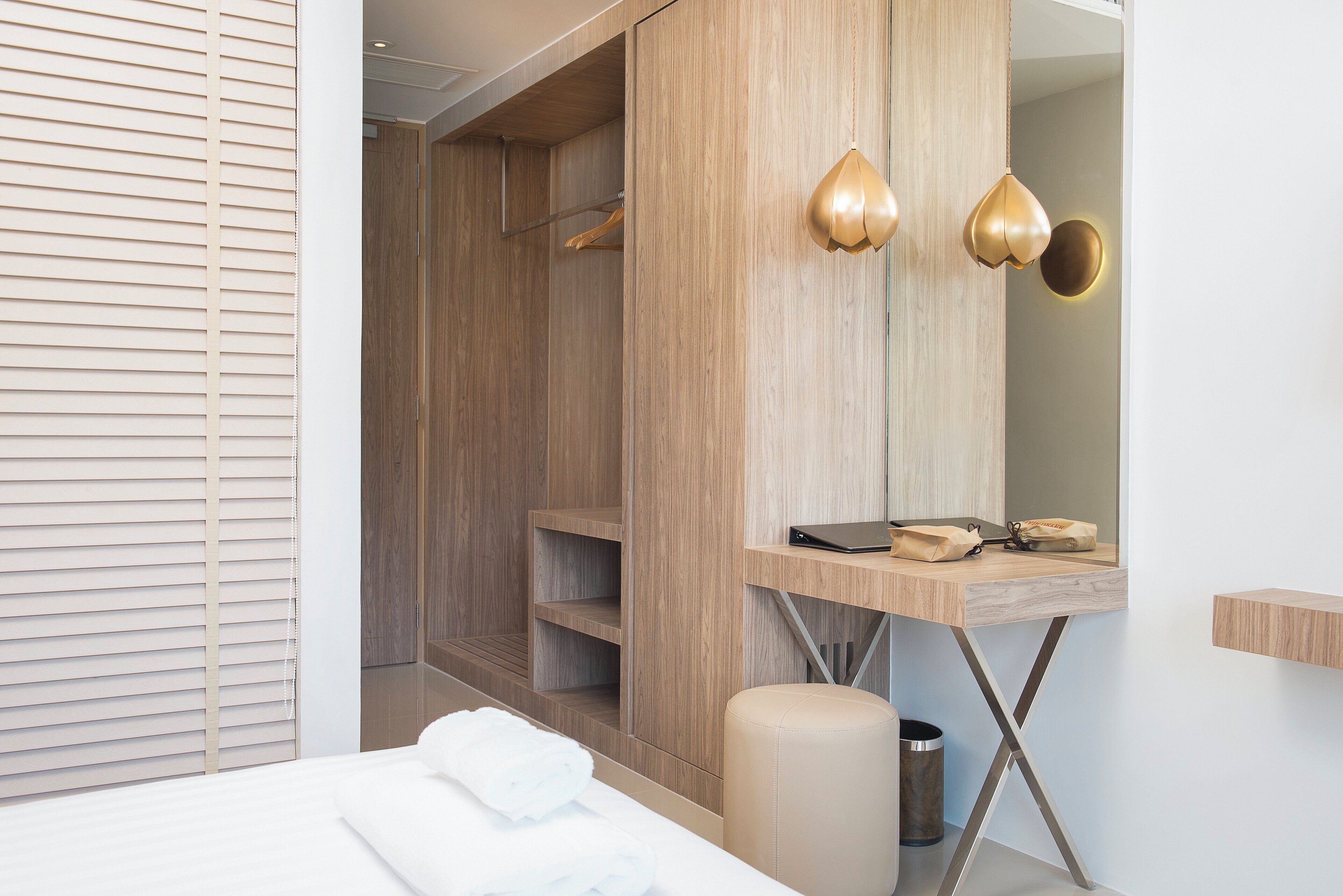 Habitación de hotel moderna con armario de madera, mesita de noche con una computadora portátil y un bolso, un cesto de basura y una cama con edredón blanco.