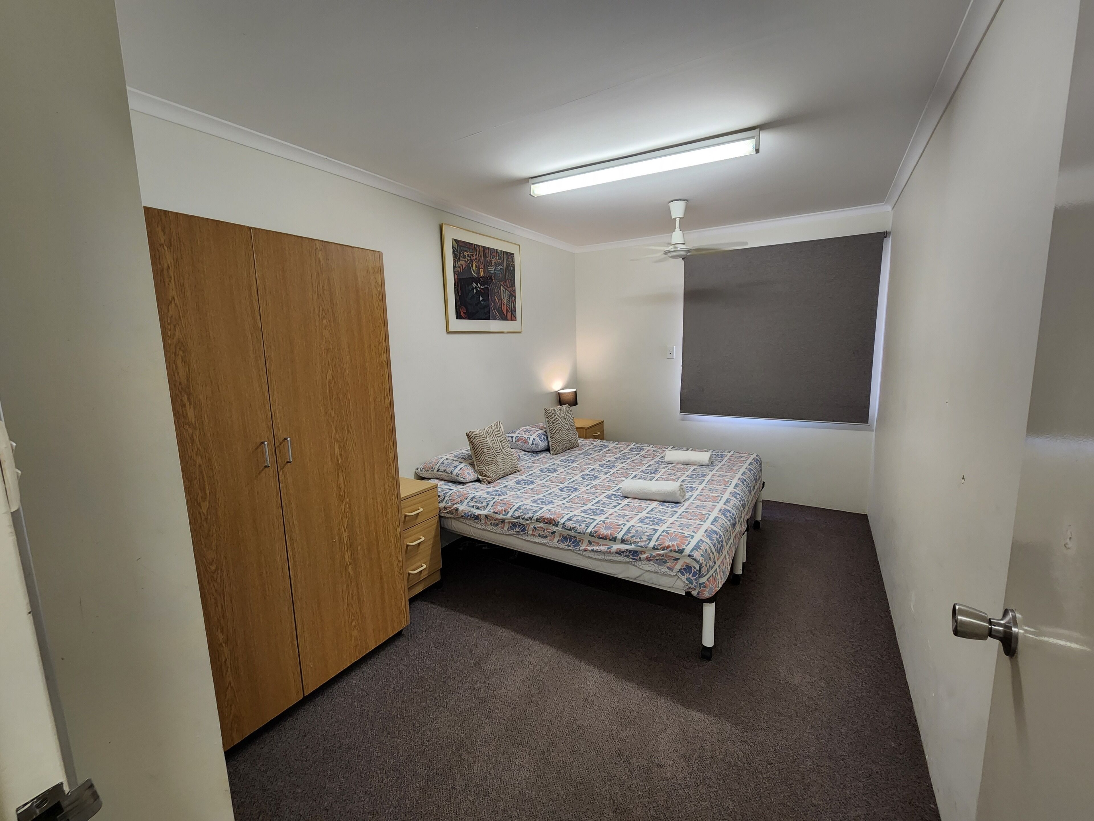 Un dormitorio con cama, armario, mesita de noche, ventilador de techo y ventana.