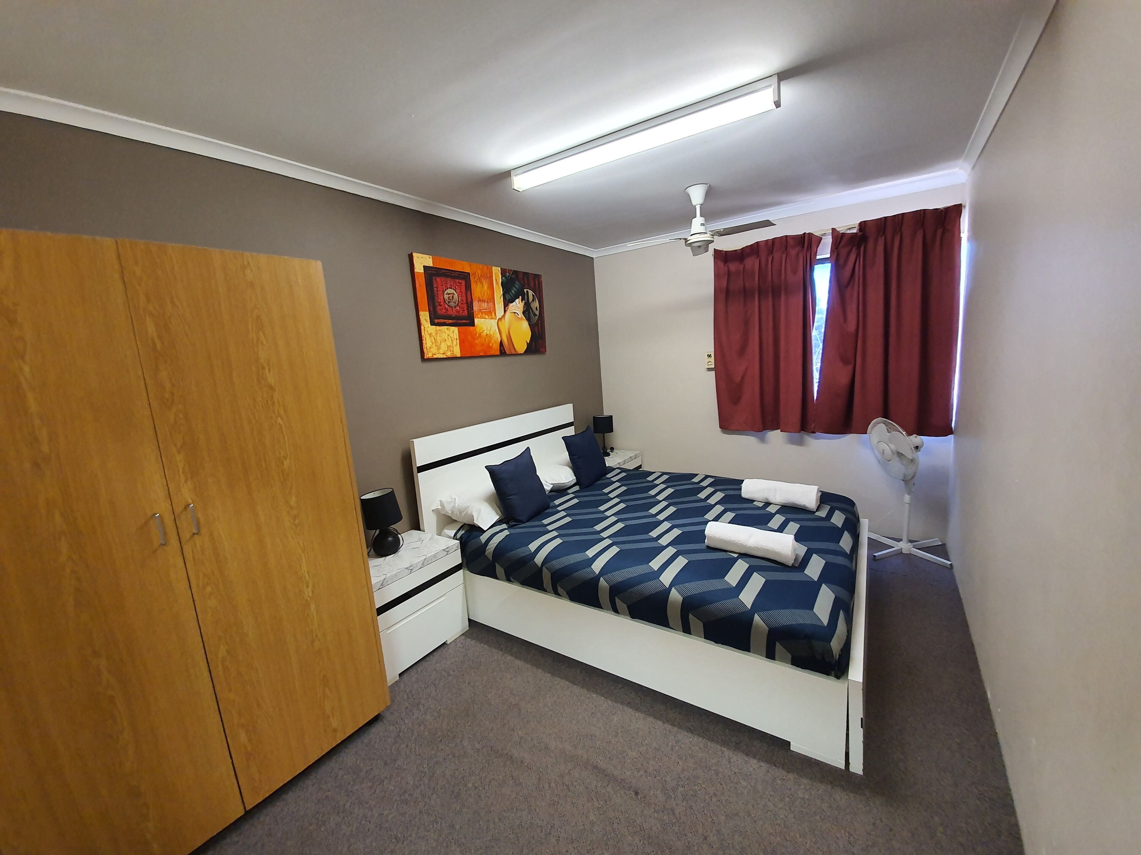 A bedroom with a bed, nightstand, wardrobe, ceiling fan, and artwork on the wall.