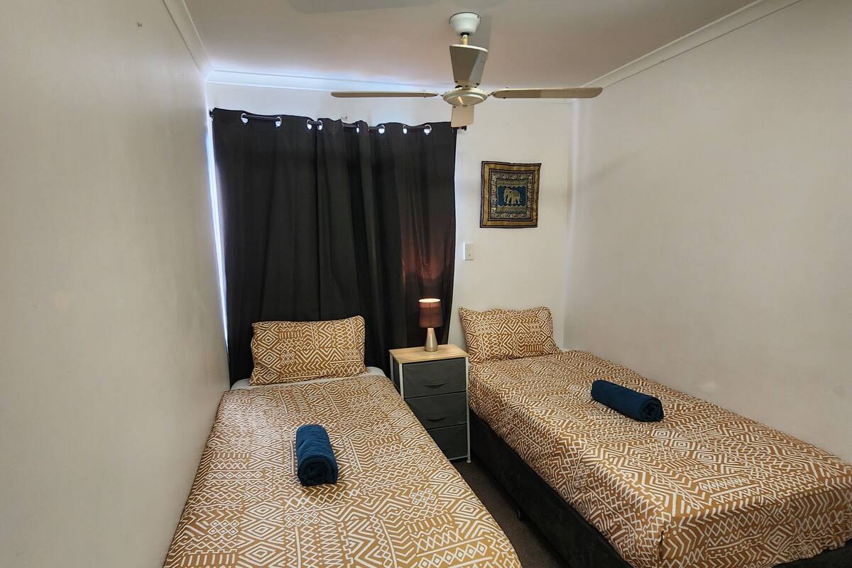 Ein Zimmer mit zwei Betten, einem Deckenventilator, einem Nachttisch mit Lampe und einem gerahmten Bild an der Wand.