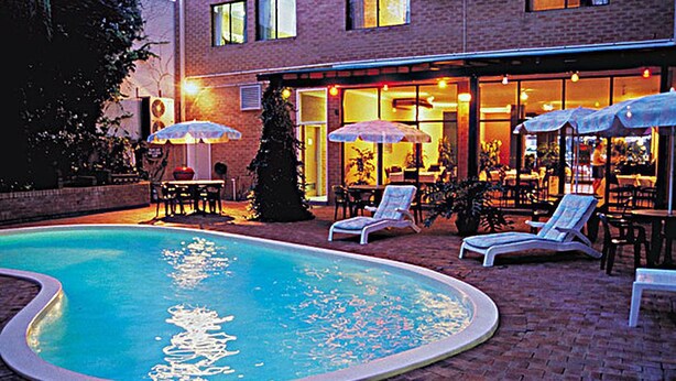 A swimming pool with lounge chairs and umbrellas, surrounded by a brick patio and a building with large windows and outdoor seating. - Perth City Apartment Hotel (West Perth)
