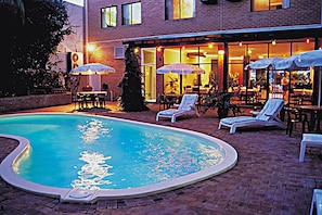 A swimming pool with lounge chairs and umbrellas, surrounded by a brick patio and a building with large windows and outdoor seating.