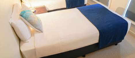 Une chambre d’hôtel avec deux lits, chacun étant équipé d’un oreiller et d’une couverture. Une lampe se trouve sur le côté gauche de la pièce.