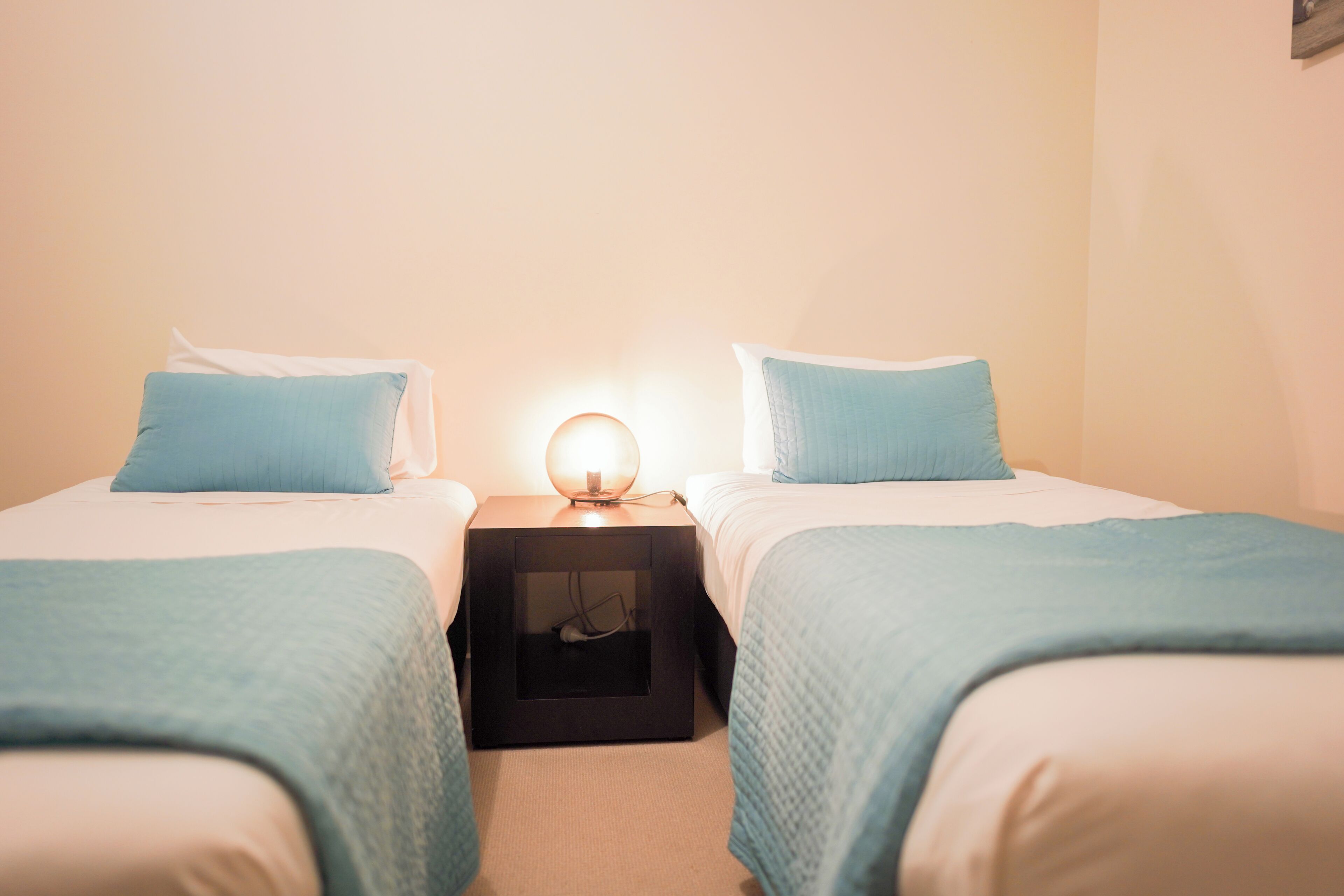 Habitación doble con dos camas, cada una con almohada y manta azules, una mesita de noche con lámpara y un pequeño mueble negro para guardar objetos.