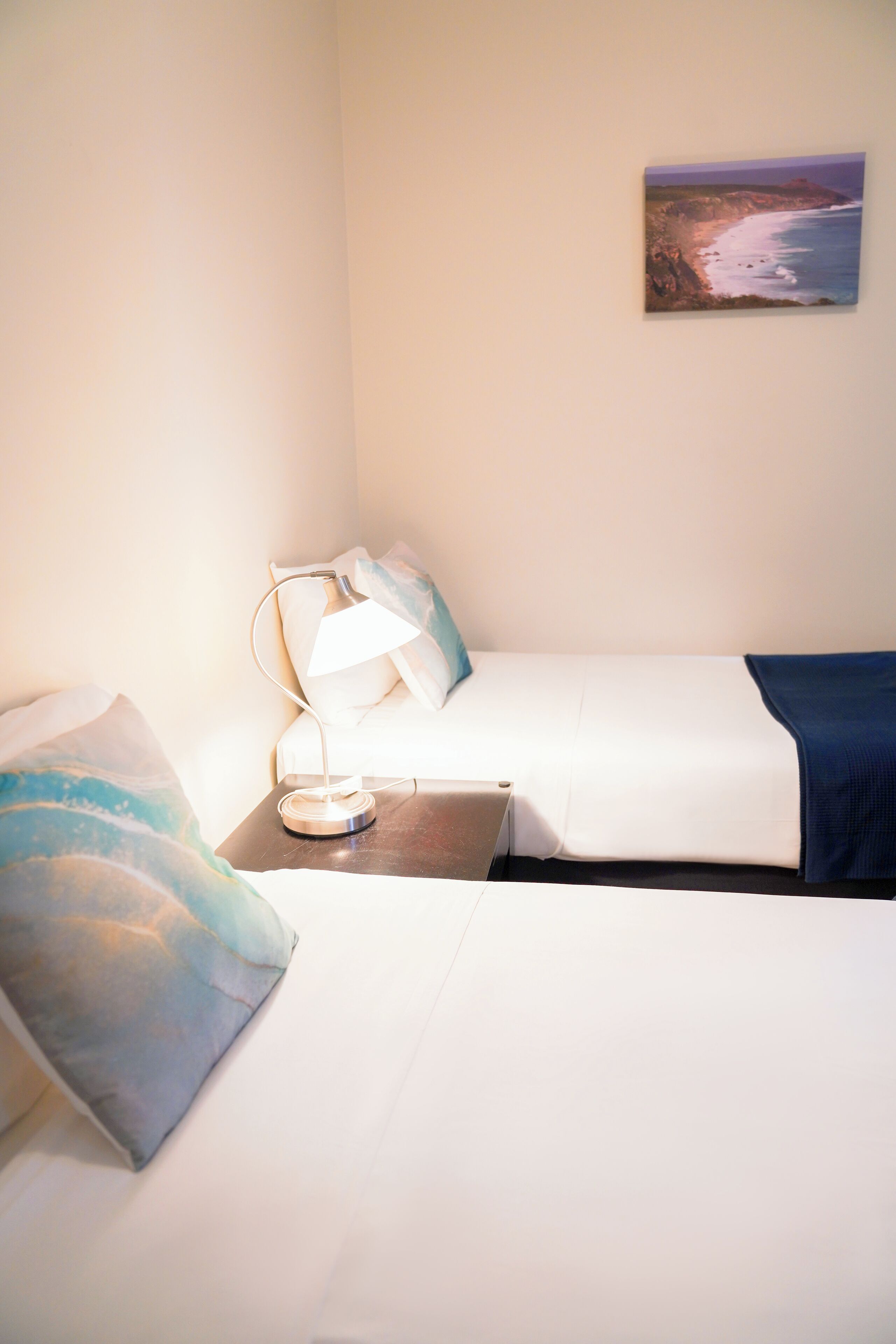 Une chambre d’hôtel avec un lit, une table de chevet avec une lampe, un oreiller et un tableau représentant un paysage côtier accroché au mur.
