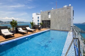 A rooftop pool with lounge chairs, a clear view of the ocean, and a modern building with a stone facade.