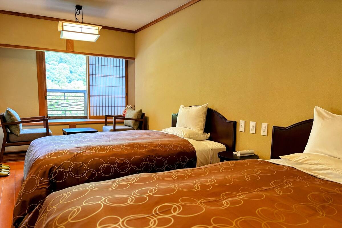 A hotel room with two beds, a desk, a chair, and a window with a view of trees.