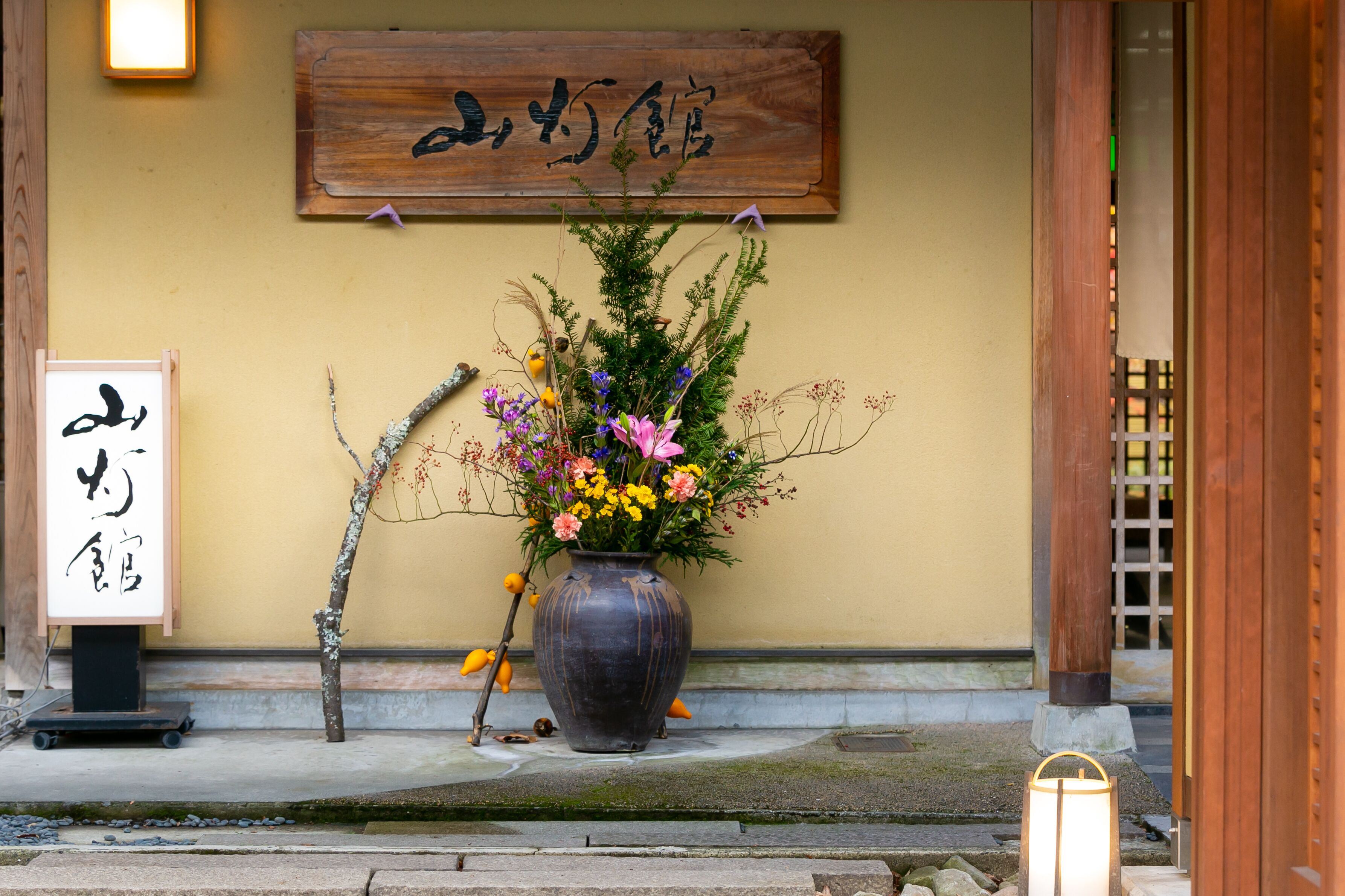 A traditional Japanese building entrance with a wooden sign, a large vase of flowers, and a lantern.