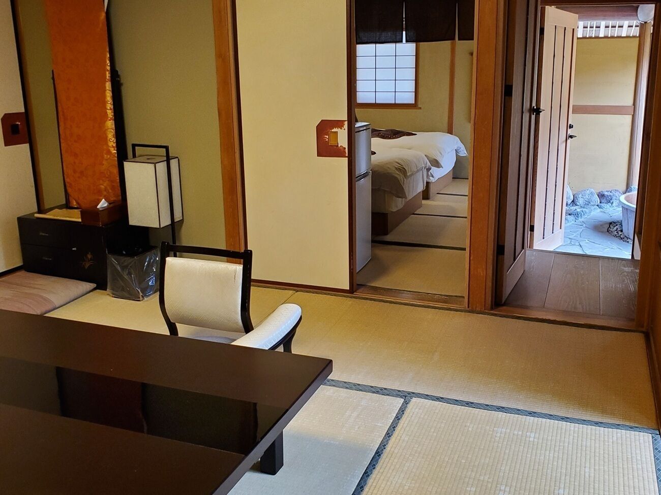A traditional Japanese-style room with tatami flooring, a low table, a chair, and a bed with a futon.