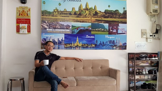 Une personne est assise sur un canapé beige dans une pièce dont un mur est décoré d’affiches de voyage et où se trouve un meuble à chaussures.