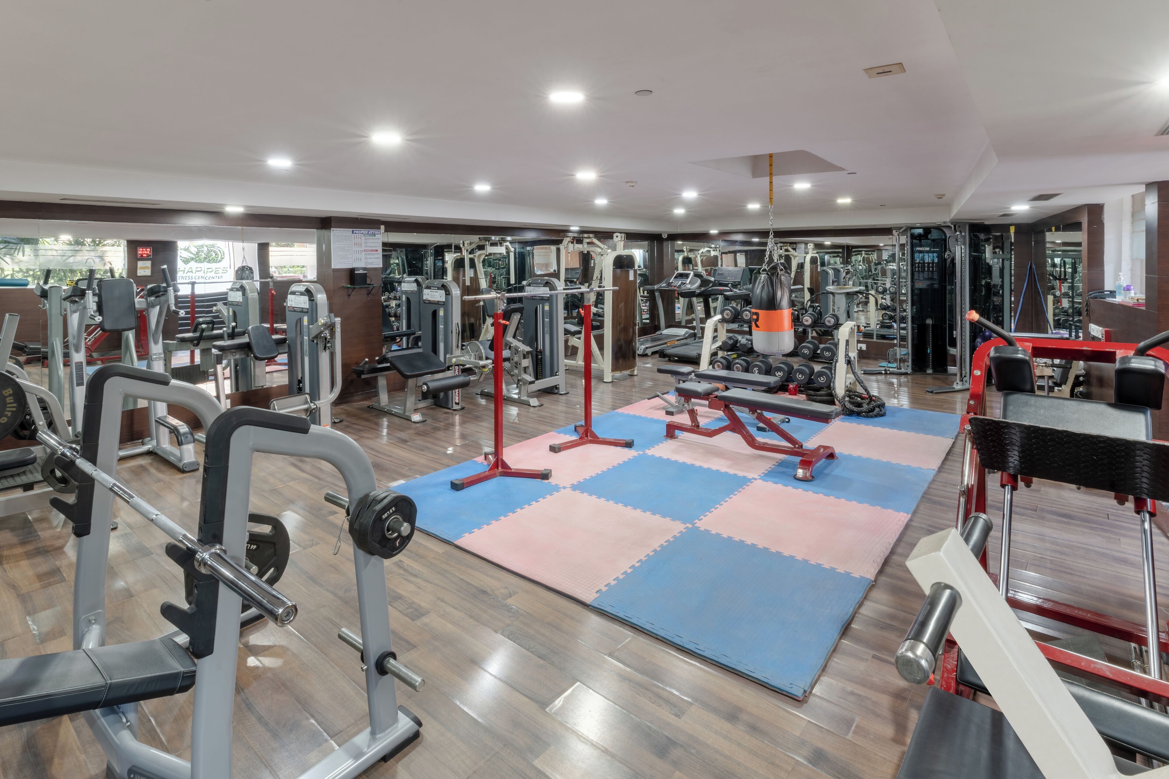 A gym with various exercise machines, a blue and pink mat area, and a wooden floor.
