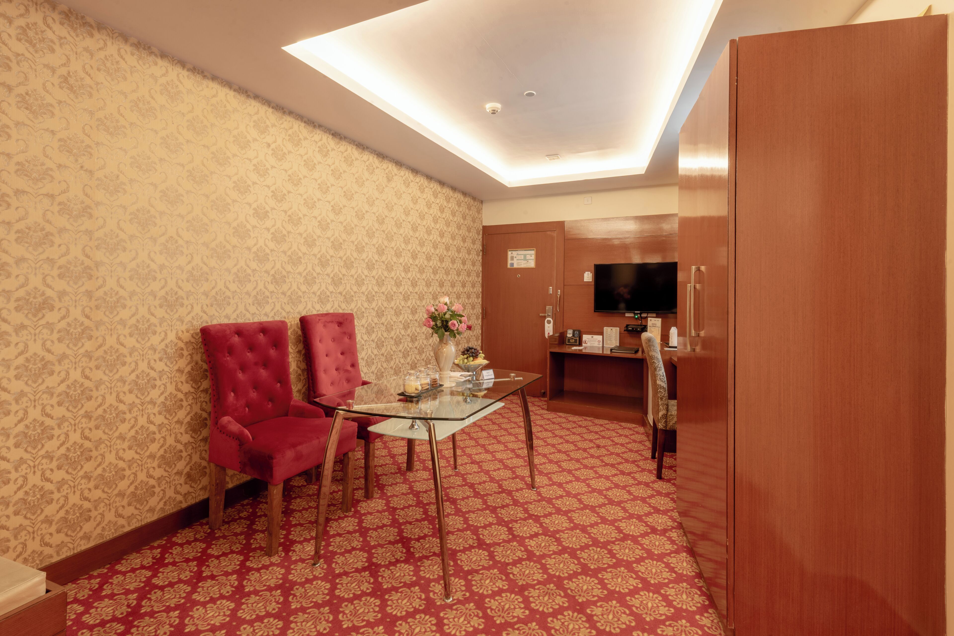 A hotel room with a patterned wallpaper, a red carpet, a glass table with chairs, a television, and a wardrobe.