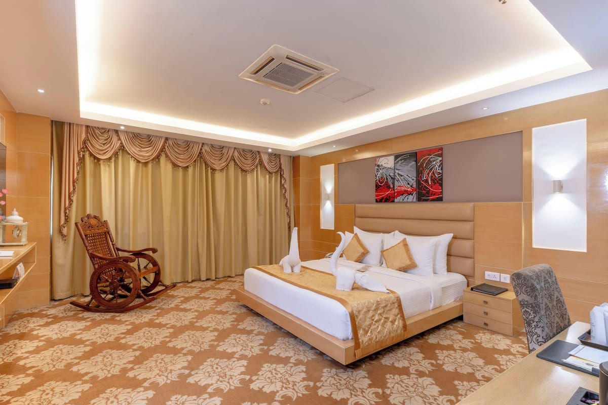 Una habitación de hotel con una cama grande, una mecedora de madera, un escritorio con una silla y un ventanal con cortinas.