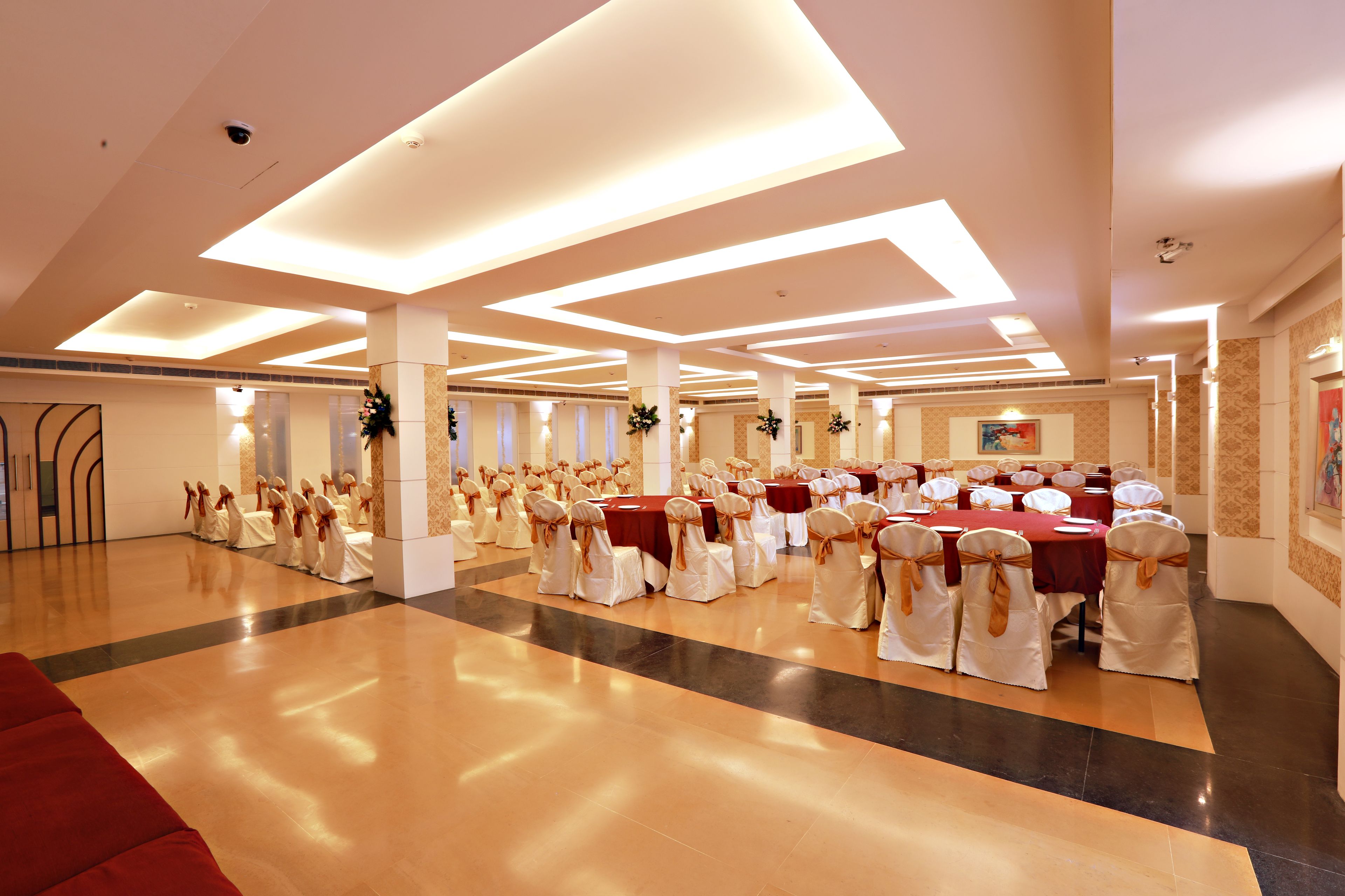 A spacious banquet hall with multiple round tables, each surrounded by white chairs with gold sashes. The room features a polished wooden floor, white walls, and a high ceiling with recessed lighting.