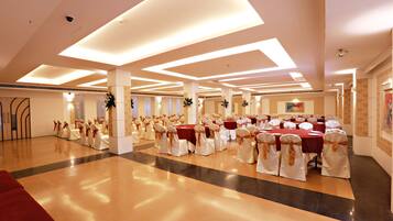 A spacious banquet hall with multiple round tables, each surrounded by white chairs with gold sashes. The room features a polished wooden floor, white walls, and a high ceiling with recessed lighting.