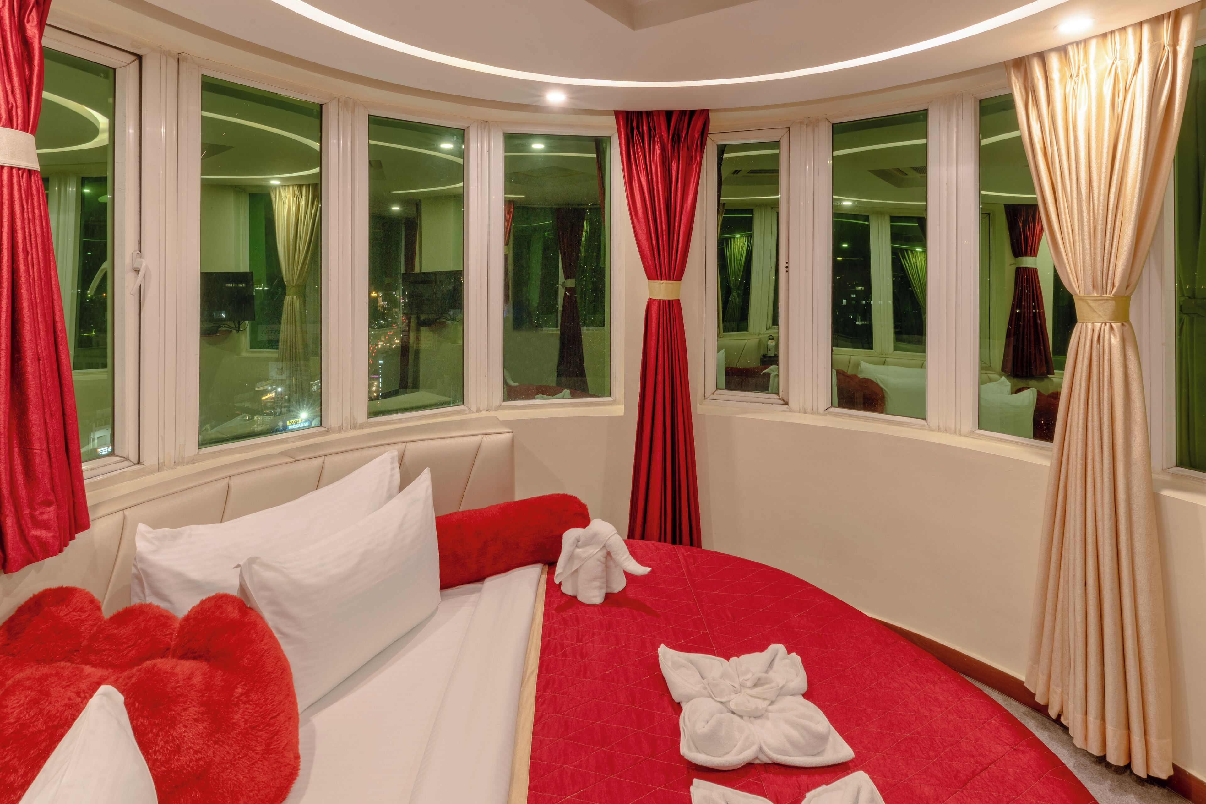 A hotel room with a large bed, red and white curtains, and multiple windows offering city views.