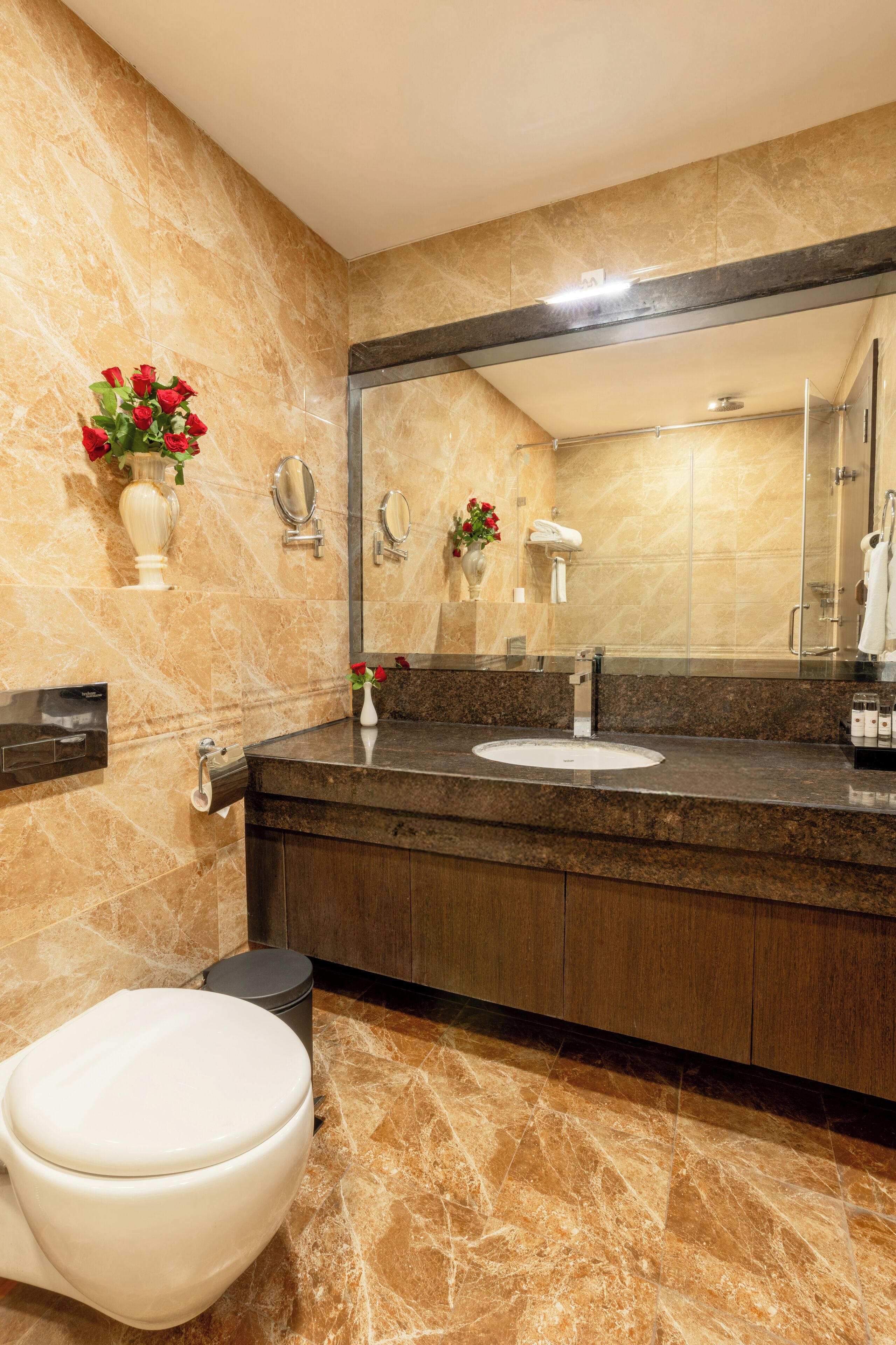 A modern bathroom with a marble floor, a white toilet, a sink with a mirror above it, and a vase with red flowers on the wall.