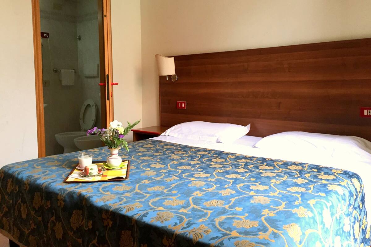 A hotel room with a bed, a bedside table with a lamp, a bathroom with a shower, and a small table with a tray of breakfast items.