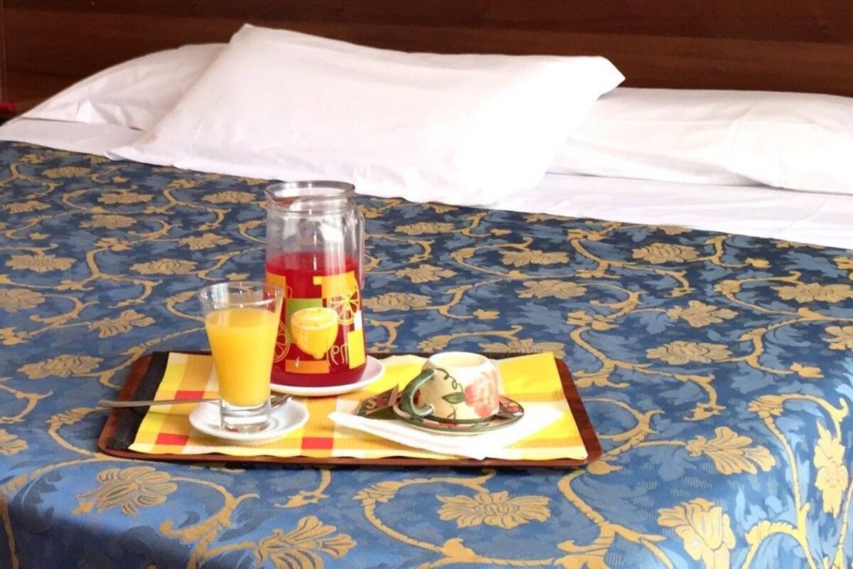 A neatly made bed with a blue patterned bedspread, a glass of orange juice, a bottle of red drink, and a cup of tea on a tray.