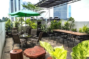 An outdoor patio with tables and chairs, surrounded by greenery and a view of city buildings.