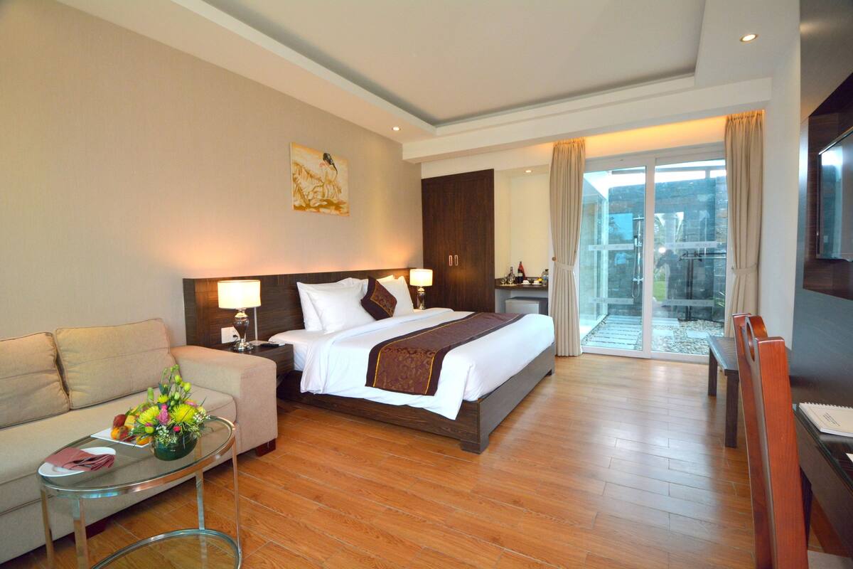 A hotel room with a bed, a sofa, a glass coffee table, and a view of the outdoors.