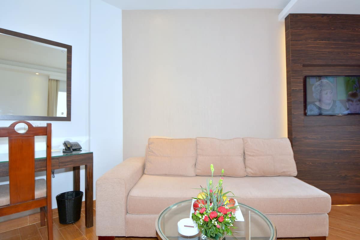A living room with a beige sofa, a glass coffee table with a vase of flowers, a wooden desk with a telephone, and a wall-mounted TV.