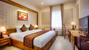 A hotel room with a large bed, a desk with a television, a chair, and a small table with a lamp.